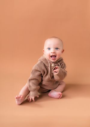 Baby in brown knit outfit smiling against peachy orange backdrop.