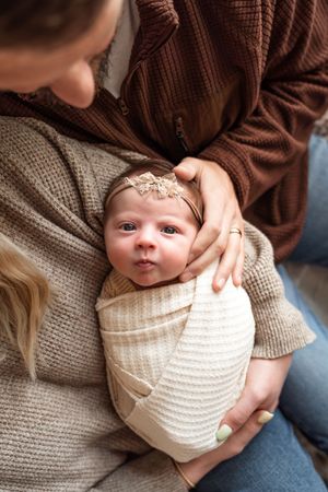 A newborn baby wrapped in a cream knit blanket wearing a delicate headband is cradled in loving arms.