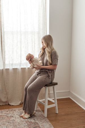 A blonde mother sits on a stool by a window, cradling her newborn baby in a soft, neutral-toned outfit.
