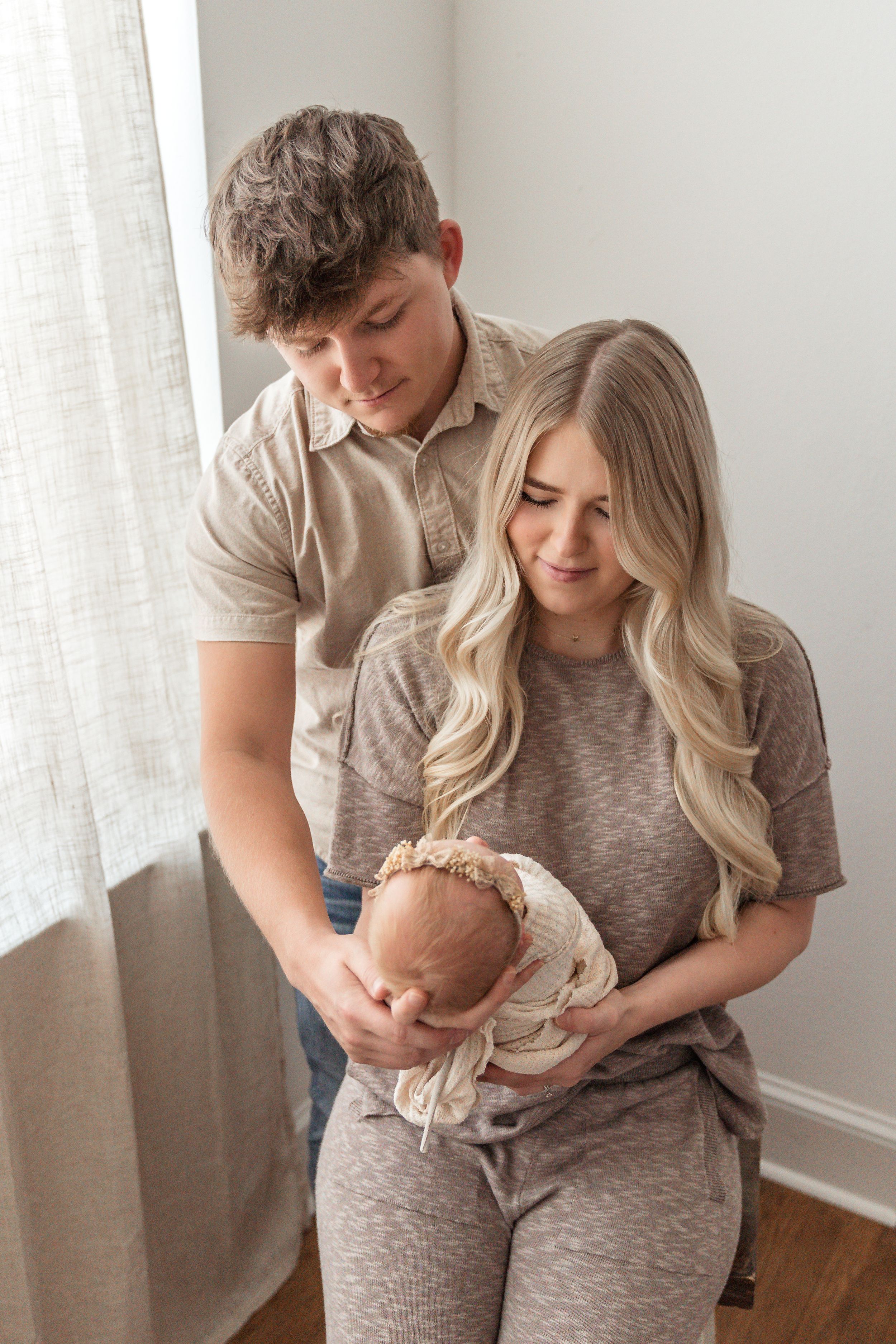 Young couple tenderly gazing at their newborn baby wrapped in a floral bonnet near a bright window.