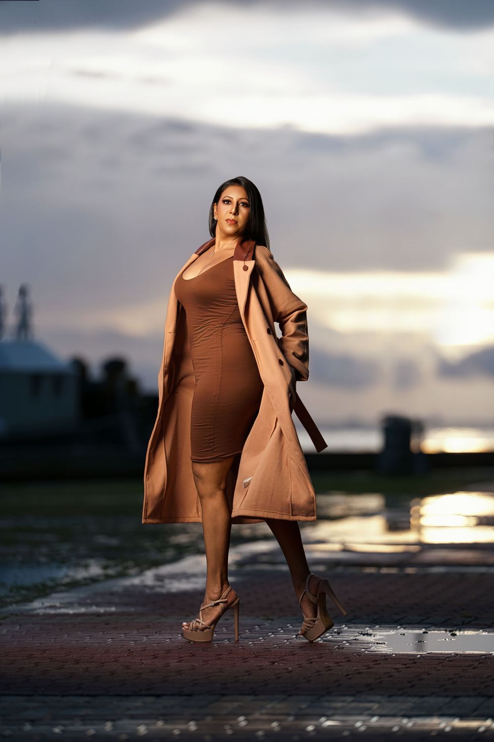 A person in a brown coat and boots poses dramatically at sunset against a reflective wet surface near a cityscape.