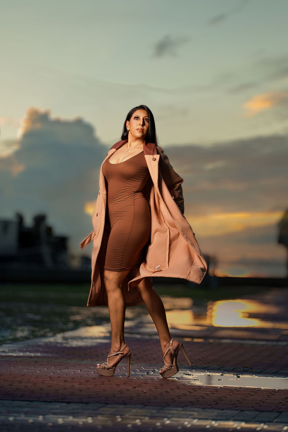 A figure in a brown bodysuit and beige coat poses gracefully on a beach at sunset with reflective water.
