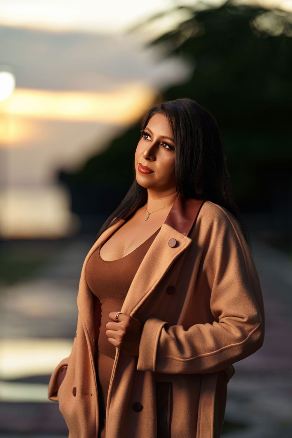 A person in a brown coat poses outdoors during golden hour with dramatic lighting and soft bokeh effect.