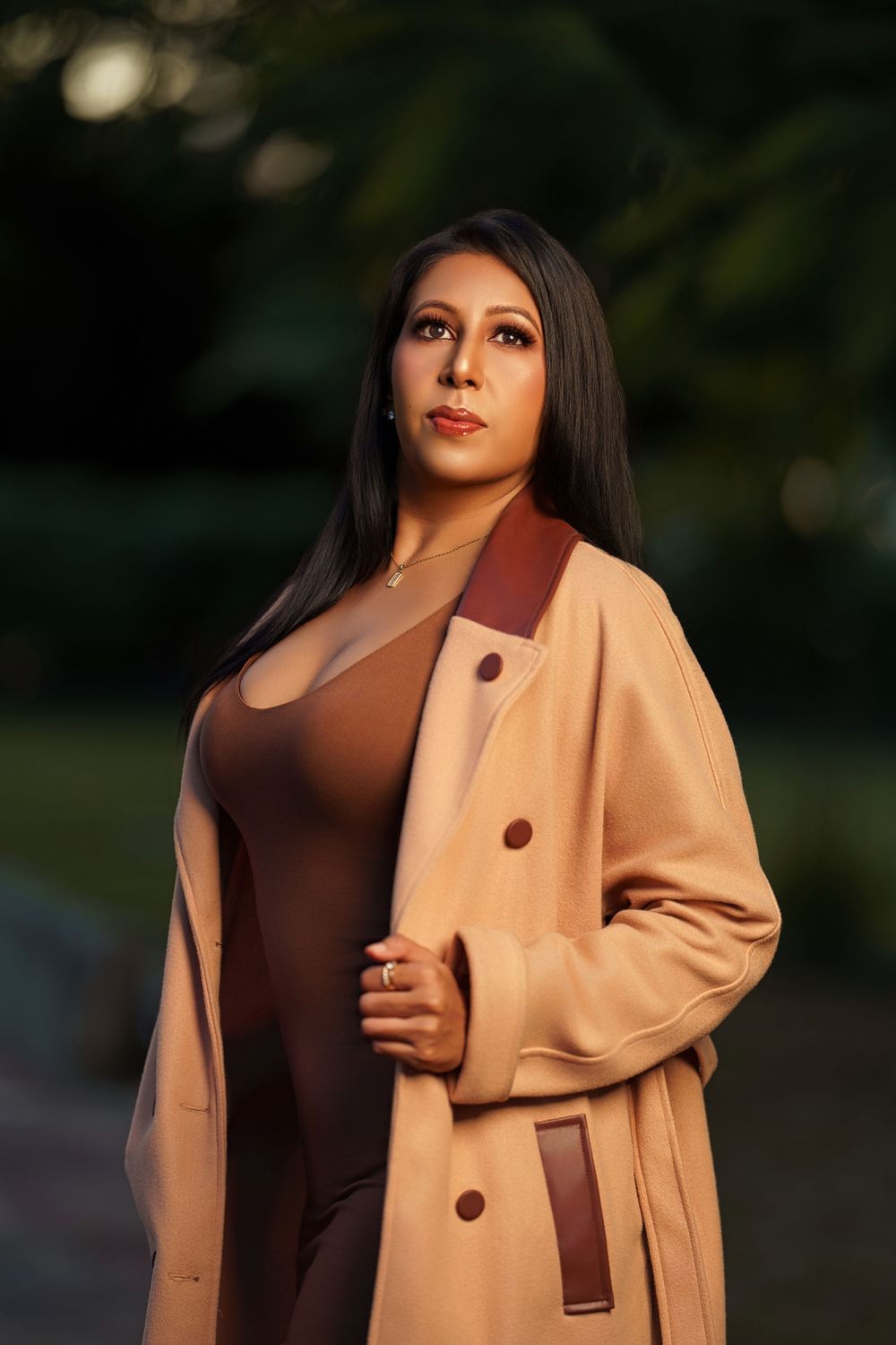 Fashion model wearing a stylish camel coat and brown dress poses elegantly in natural outdoor lighting.