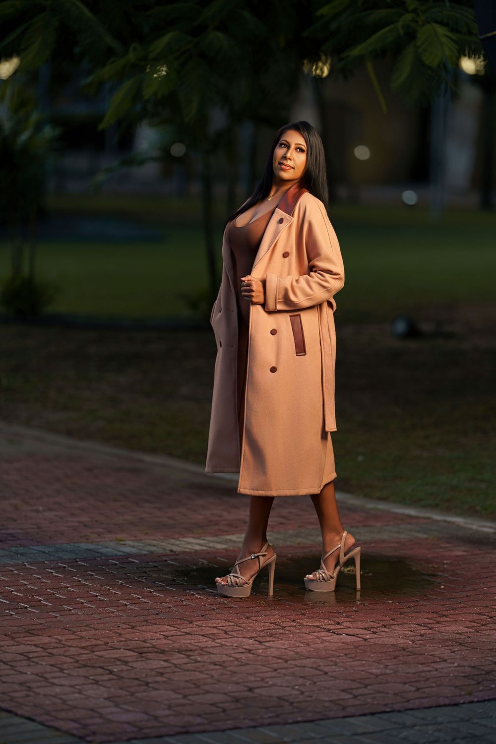 Woman in peach coat and brown pants poses stylishly on brick pathway during golden hour lighting.