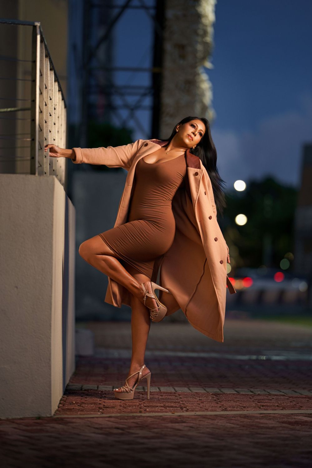 Fashion model in brown bodycon dress and camel coat poses dramatically against wall at night with city lights in background.