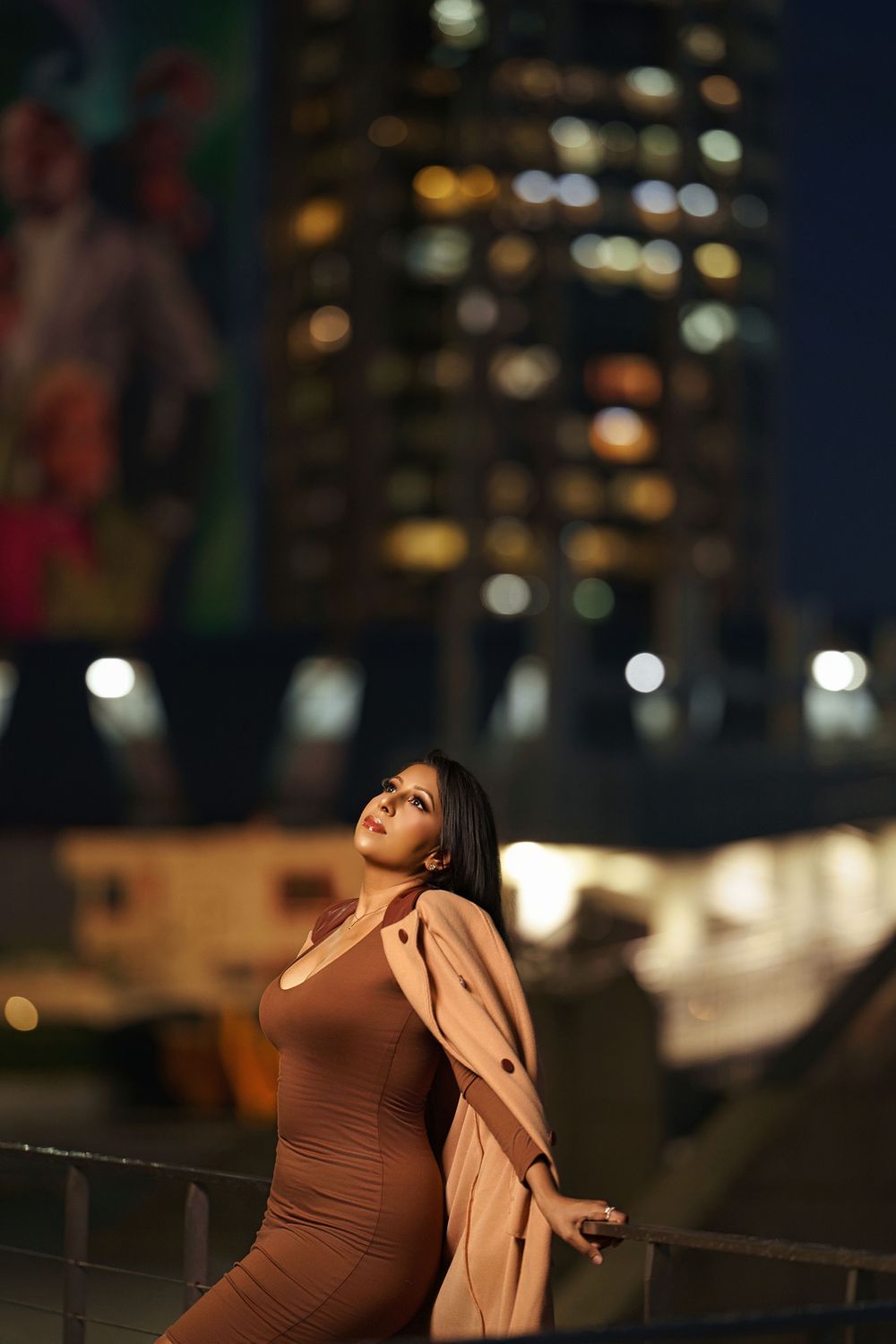 Evening fashion shoot featuring a brown dress and light coat against a cityscape with twinkling lights.