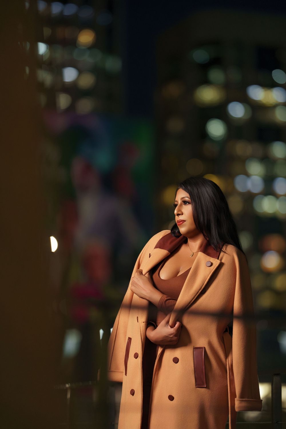 A woman in a beige coat poses against a blurred city skyline with bokeh lights in the background.