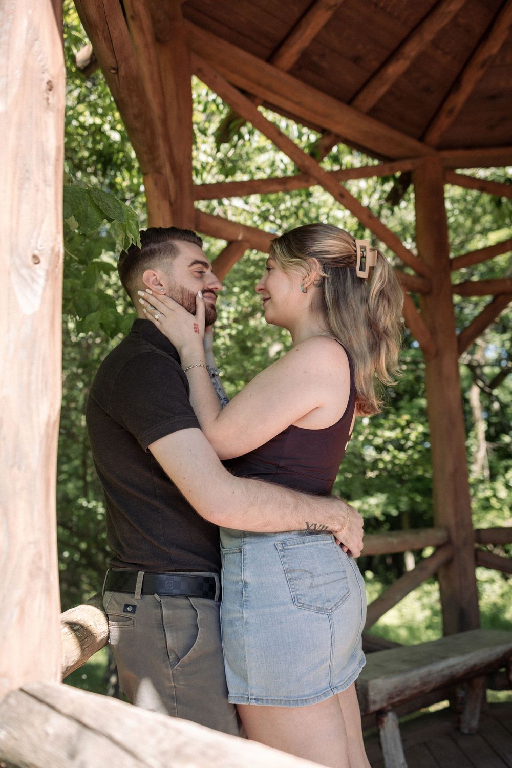 A couple shares a romantic moment under a wooden gazebo surrounded by lush green trees.
