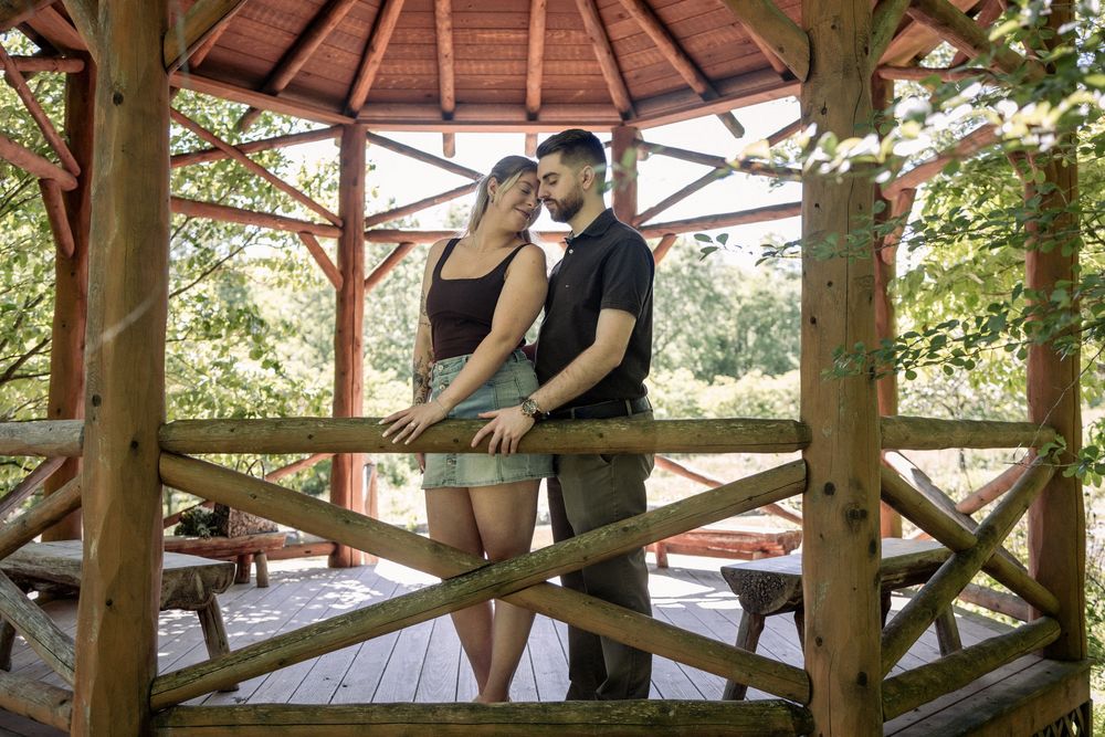A couple shares a romantic moment in a wooden gazebo surrounded by lush green trees on a sunny day.