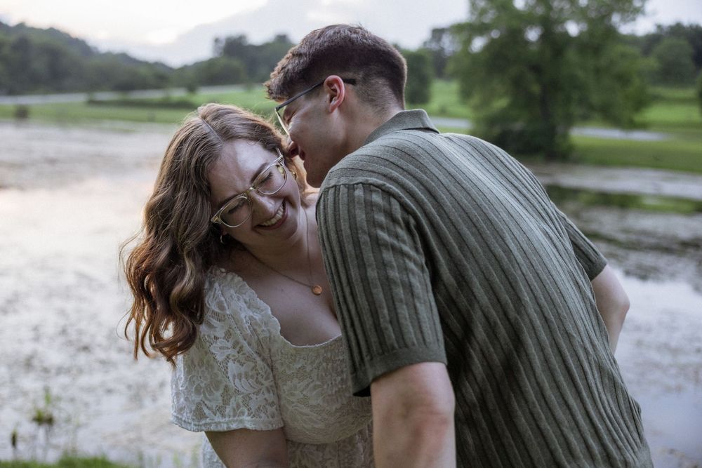 A young couple shares a tender moment outdoors near a misty lake, the man kissing the laughing woman's cheek.