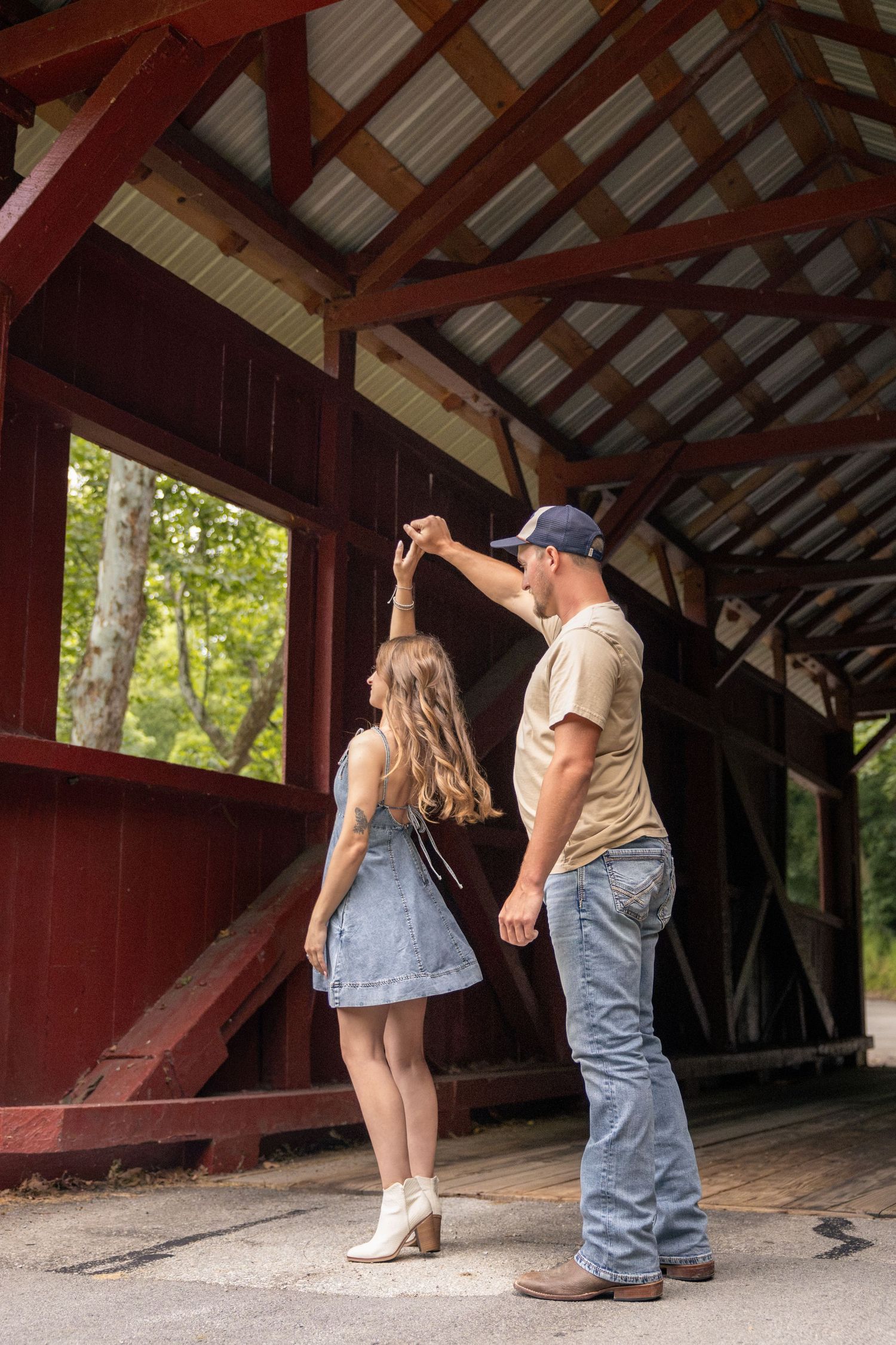 A couple dancing in a rustic red covered bridge, man spinning woman in denim dress and white boots.