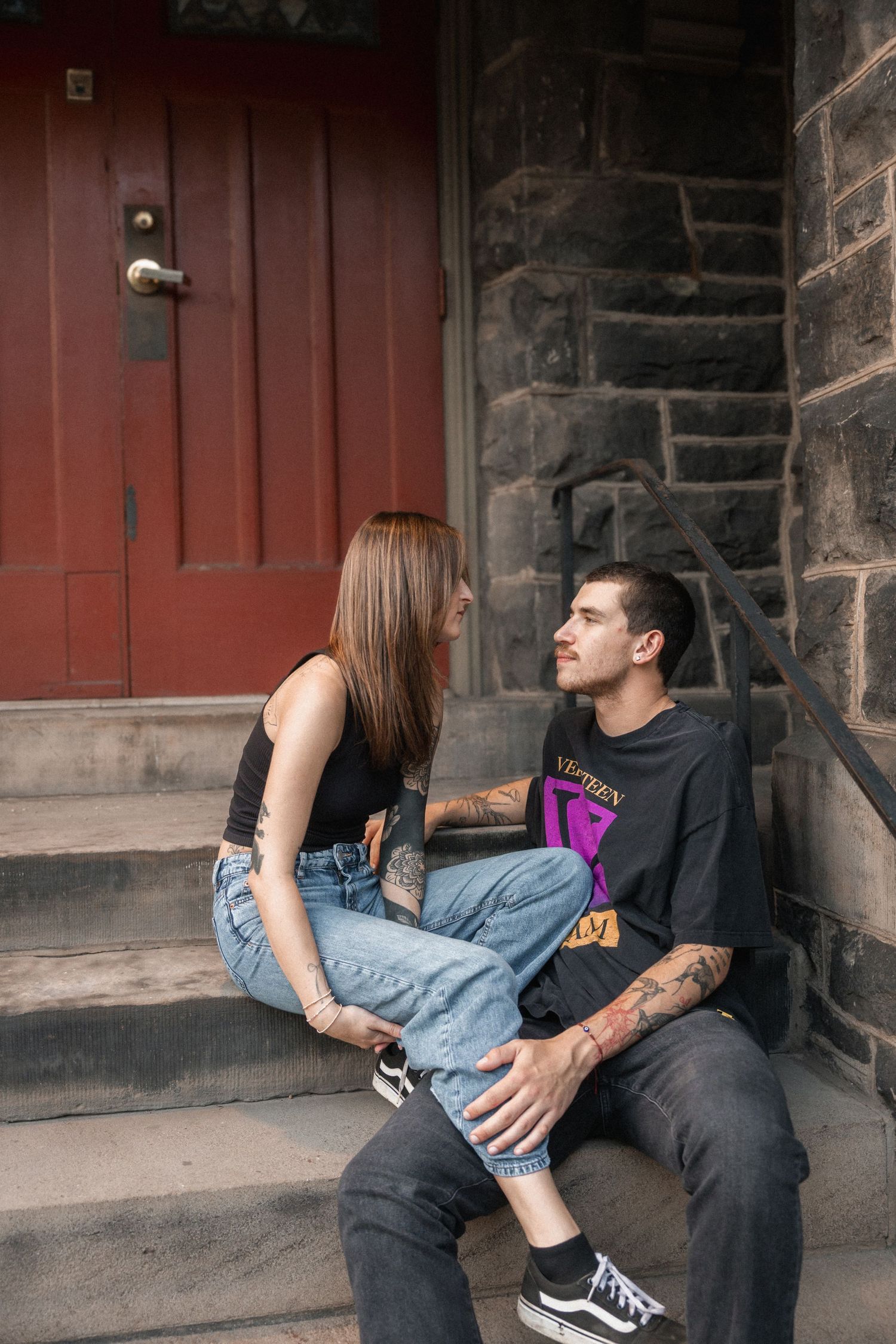 Tattooed couple sitting on stone steps by a red door, sharing an intimate moment in casual streetwear.