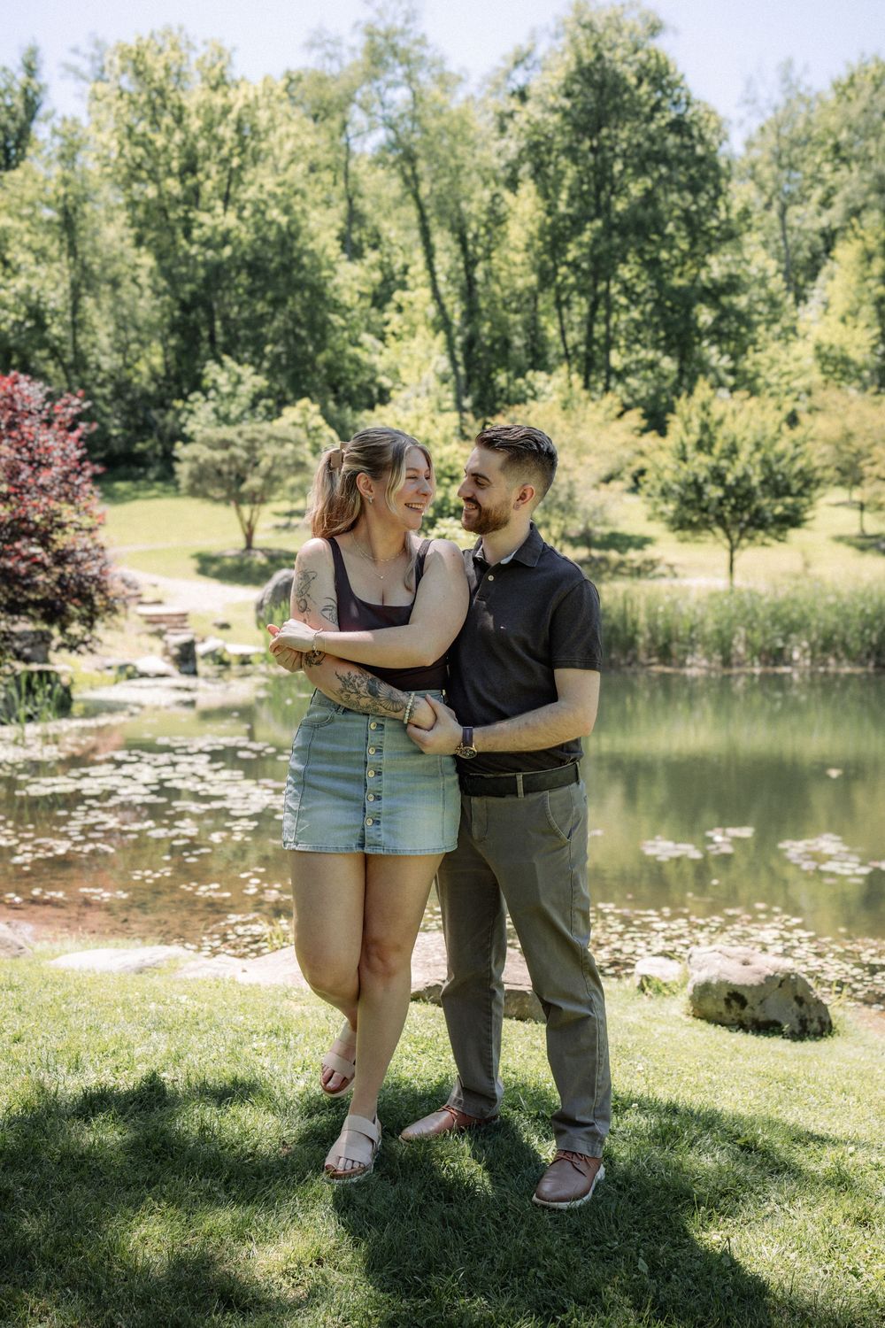Happy couple embracing and smiling at each other in a lush green park with a serene pond backdrop.