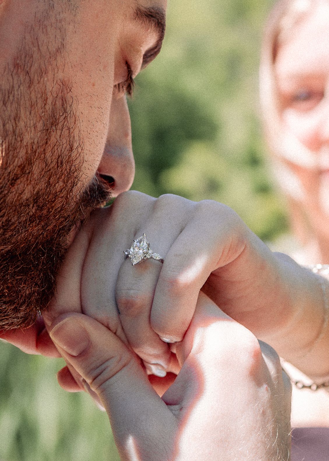 Bearded man kissing woman's hand featuring a sparkling marquise diamond engagement ring outdoors.