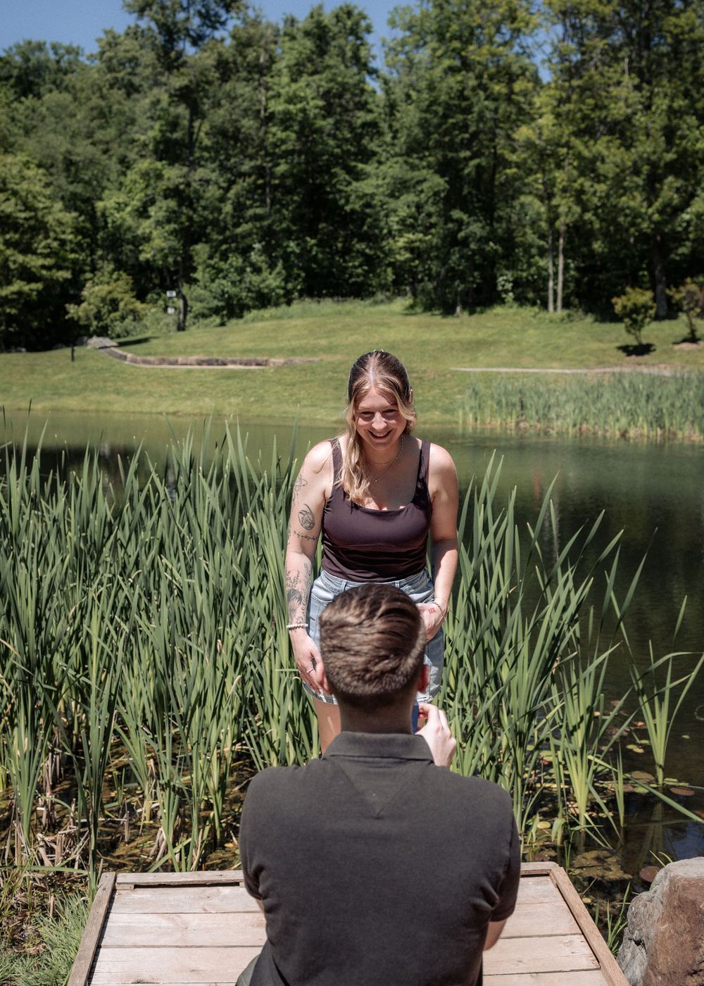 A man kneeling on a dock proposing to a smiling woman near a serene pond surrounded by lush green trees.