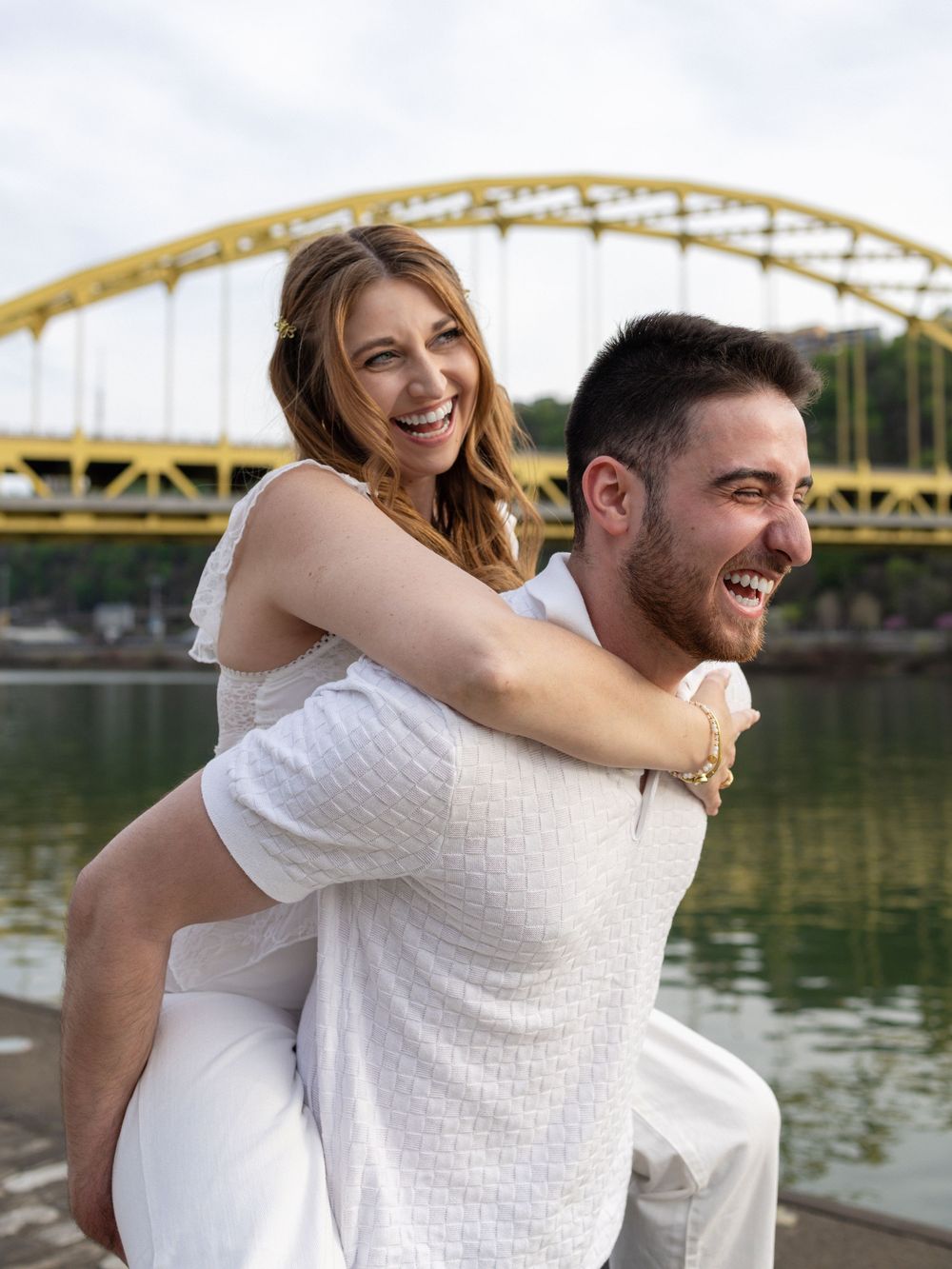 Happy couple in white outfits laughing during a piggyback ride near a yellow bridge by the river.