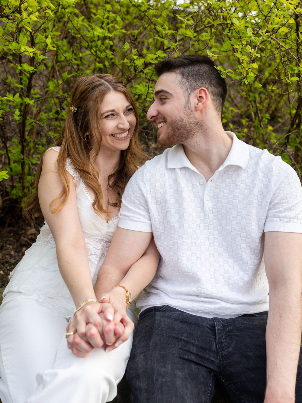Happy couple holding hands and smiling at each other outdoors, surrounded by lush green spring foliage.