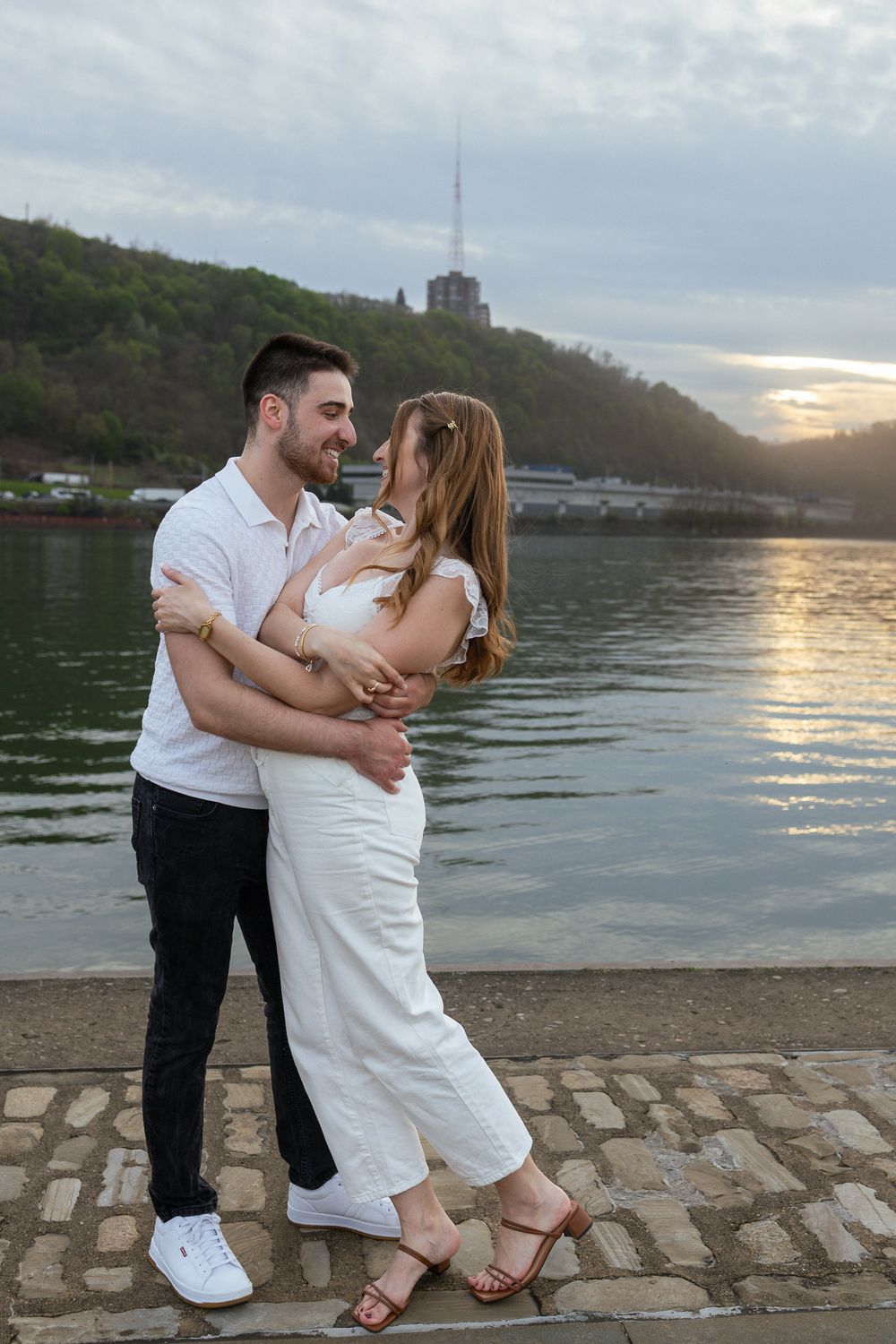 Couple embracing and smiling at each other by a riverside at sunset, both wearing white outfits.