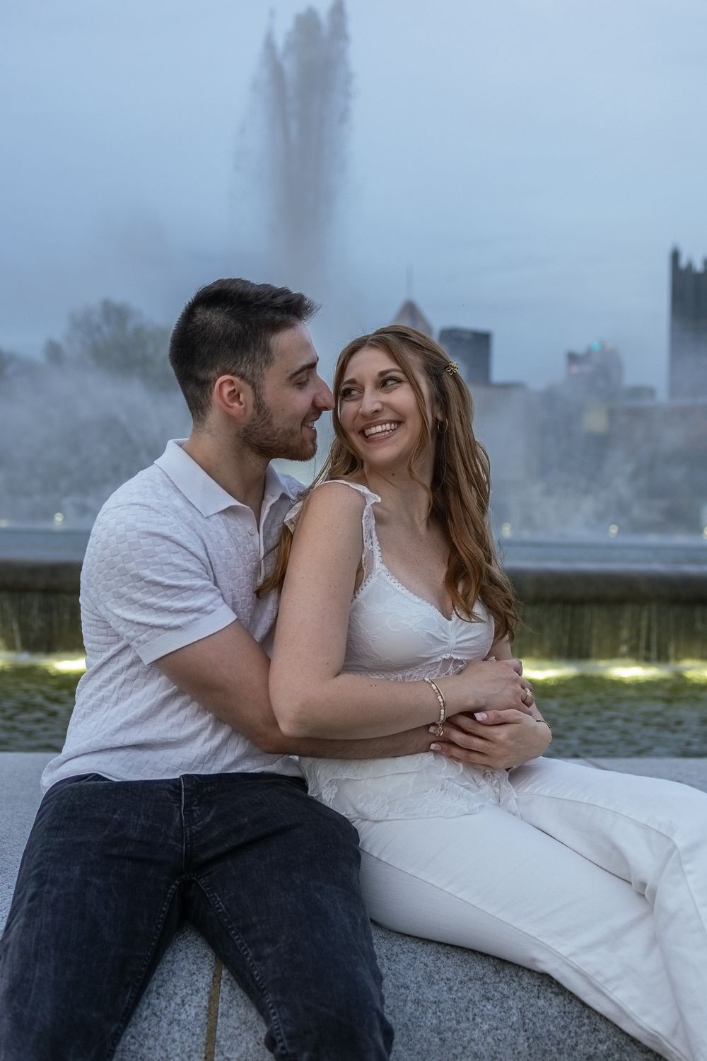 Romantic couple sitting together by a fountain at dusk, woman in white dress smiling at partner.