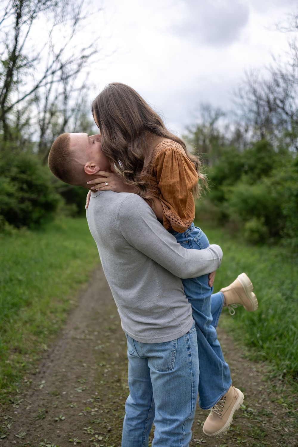 A man lifts and kisses a woman on a forest path, both wearing jeans in a romantic outdoor embrace.
