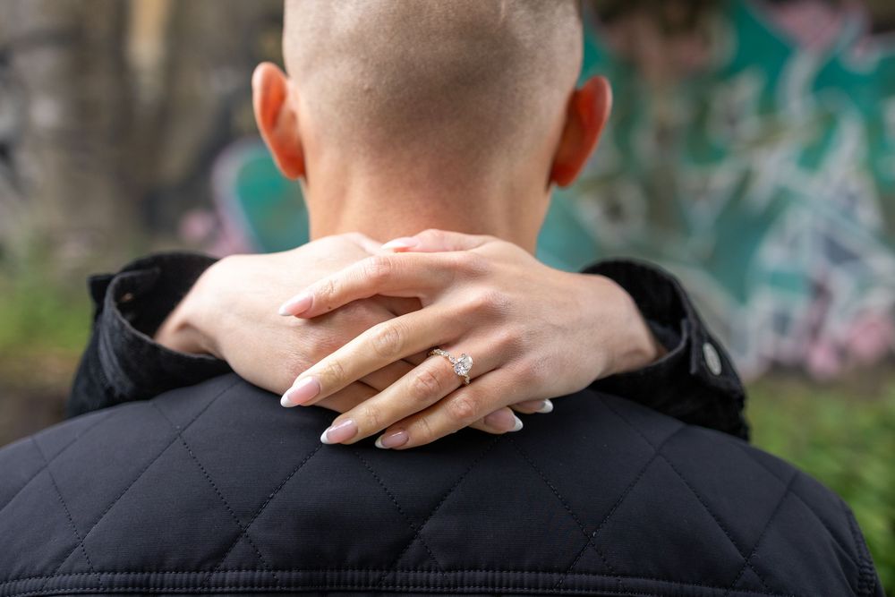 Woman with manicured nails and engagement ring resting hands on shaved head partner wearing black jacket.