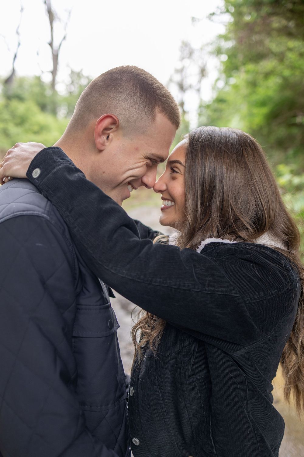 A young couple smiles and touches foreheads affectionately outdoors, wearing dark jackets with green trees in the background.