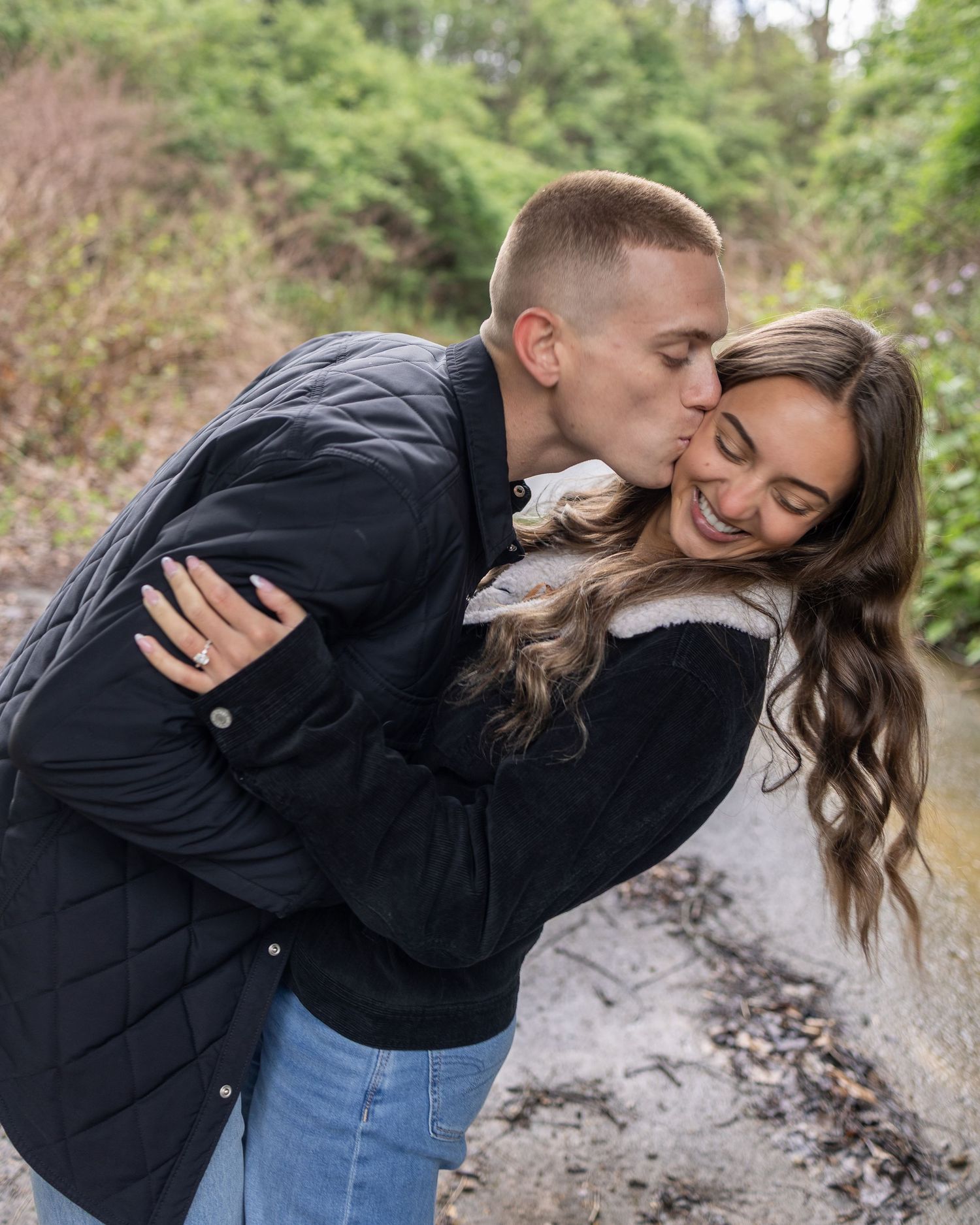 Couple sharing a romantic moment outdoors, man kissing woman on cheek, both wearing dark jackets, engagement ring visible.