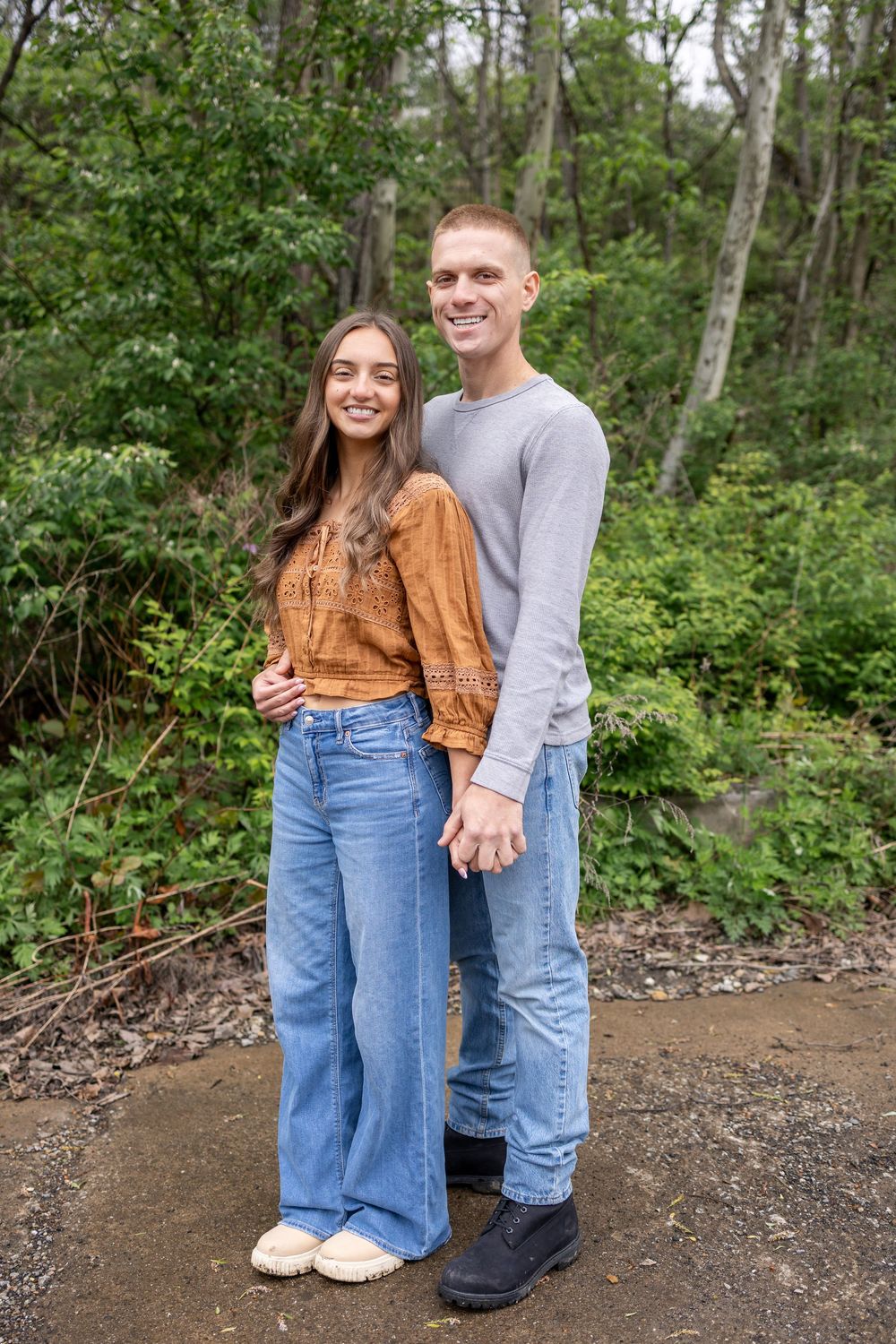 A smiling couple poses together on a forest path, wearing jeans and casual tops, surrounded by lush green trees.