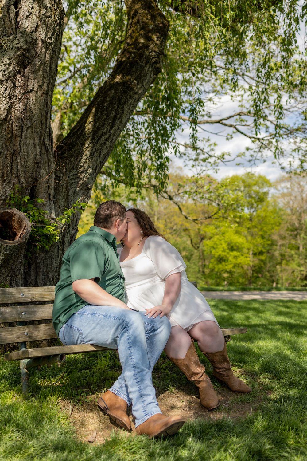 Couple sharing a romantic kiss on a wooden bench under a willow tree in a lush green park.