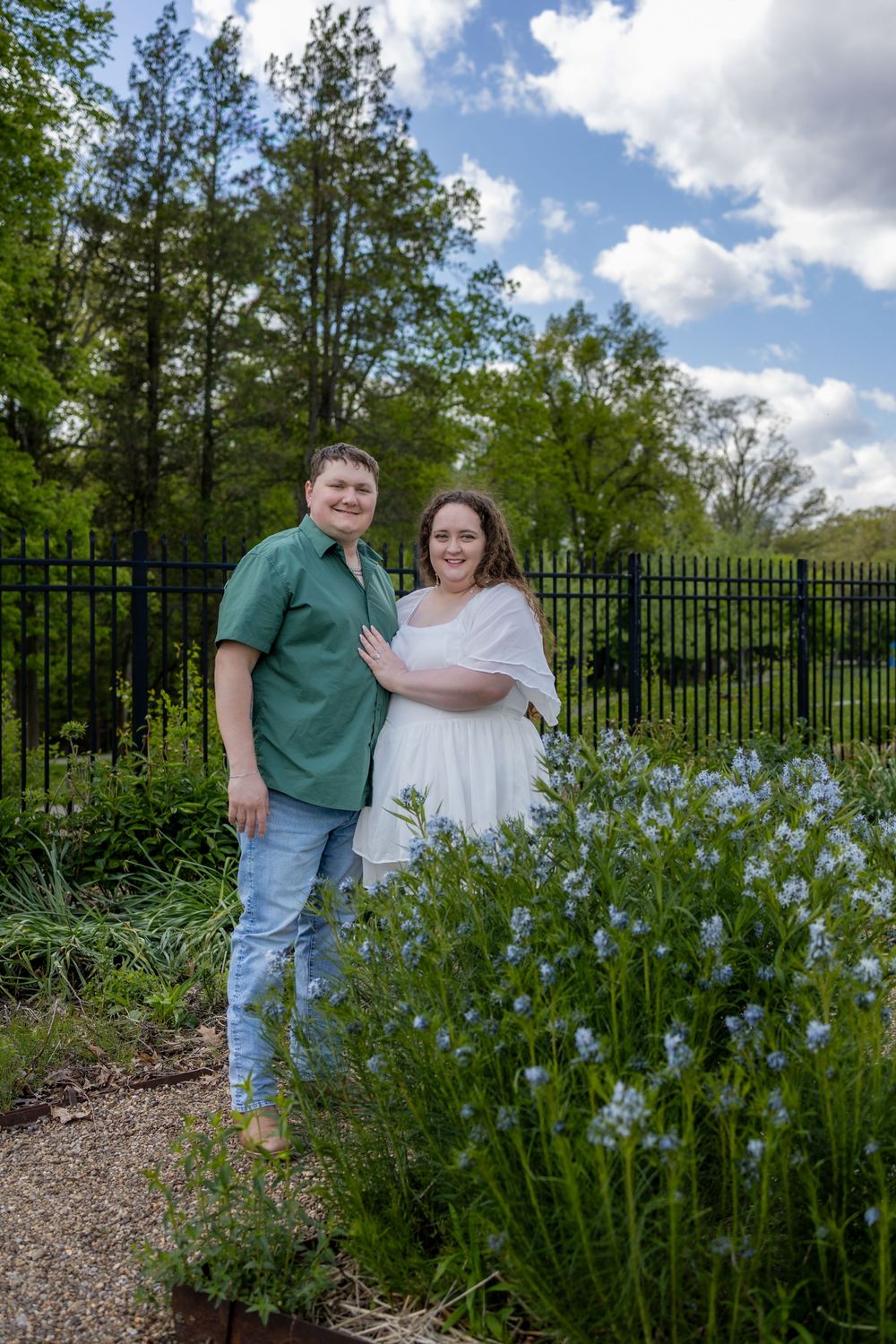 Smiling couple posing together in a garden with blue flowers, green fence, and cloudy sky.