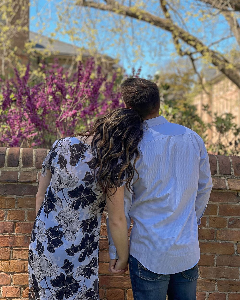 Couple standing together facing a brick wall with blooming purple trees and spring foliage in the background.