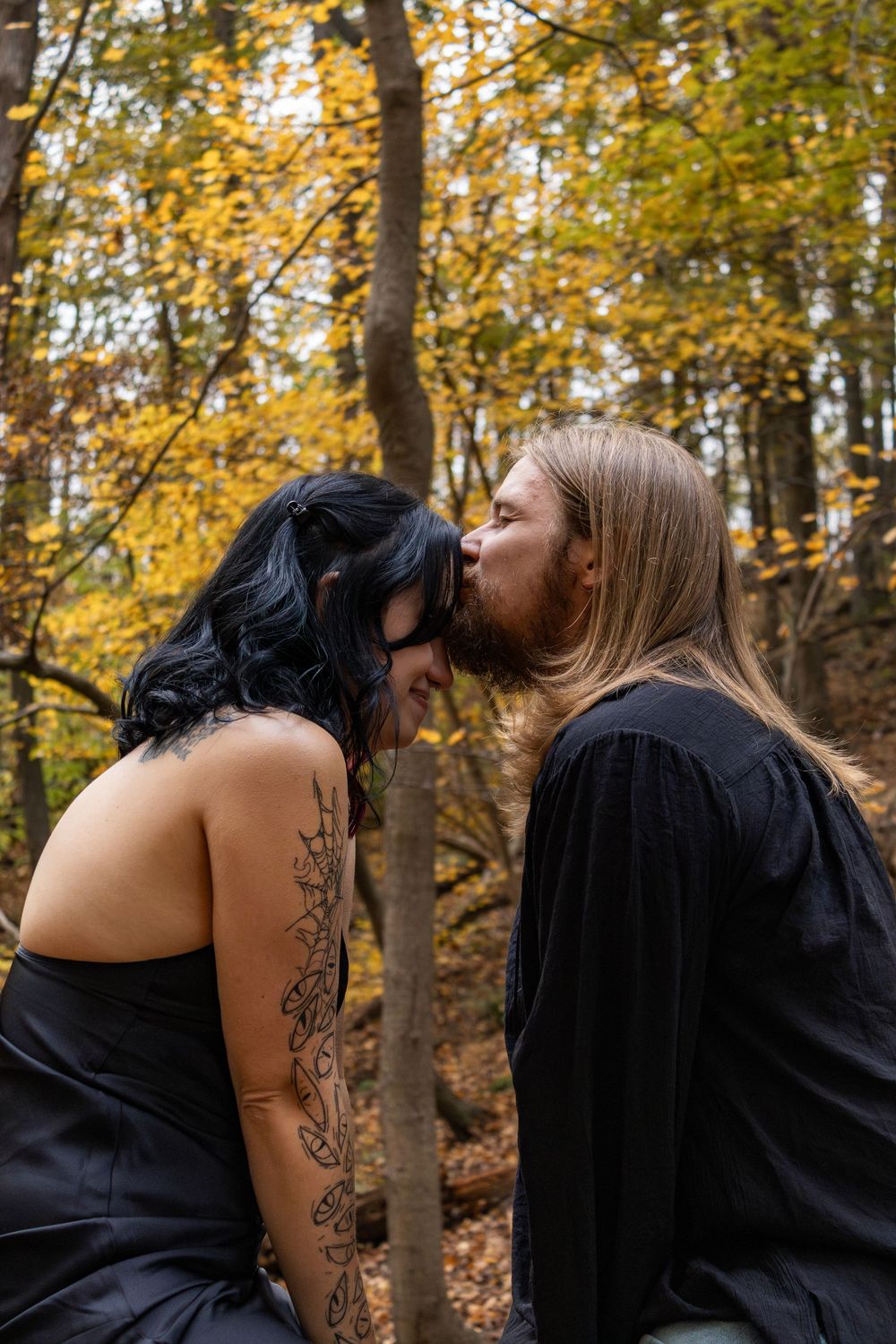 Couple sharing a kiss in autumn forest, woman with black hair and floral arm tattoo, man with long hair, both in black.