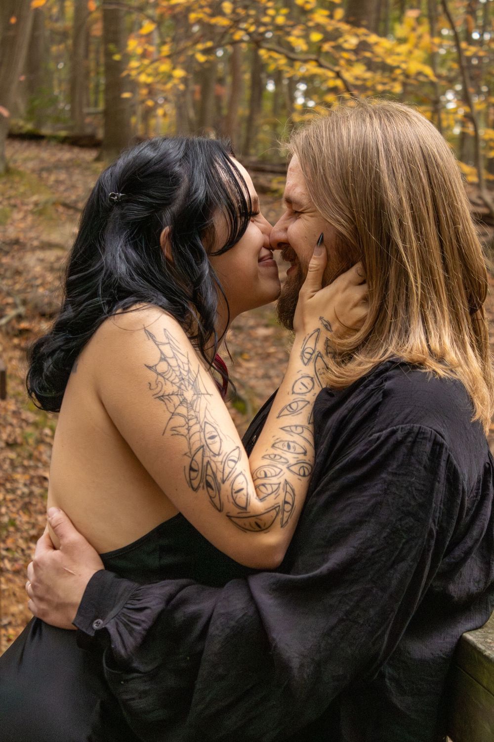 Two women with tattoos share a romantic moment in an autumn forest, wearing black outfits.