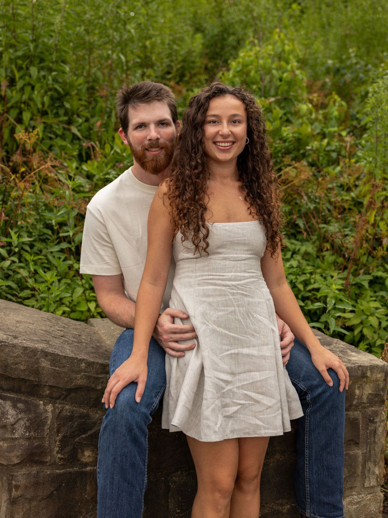 A smiling couple poses outdoors near a stone wall with lush green foliage in the background.