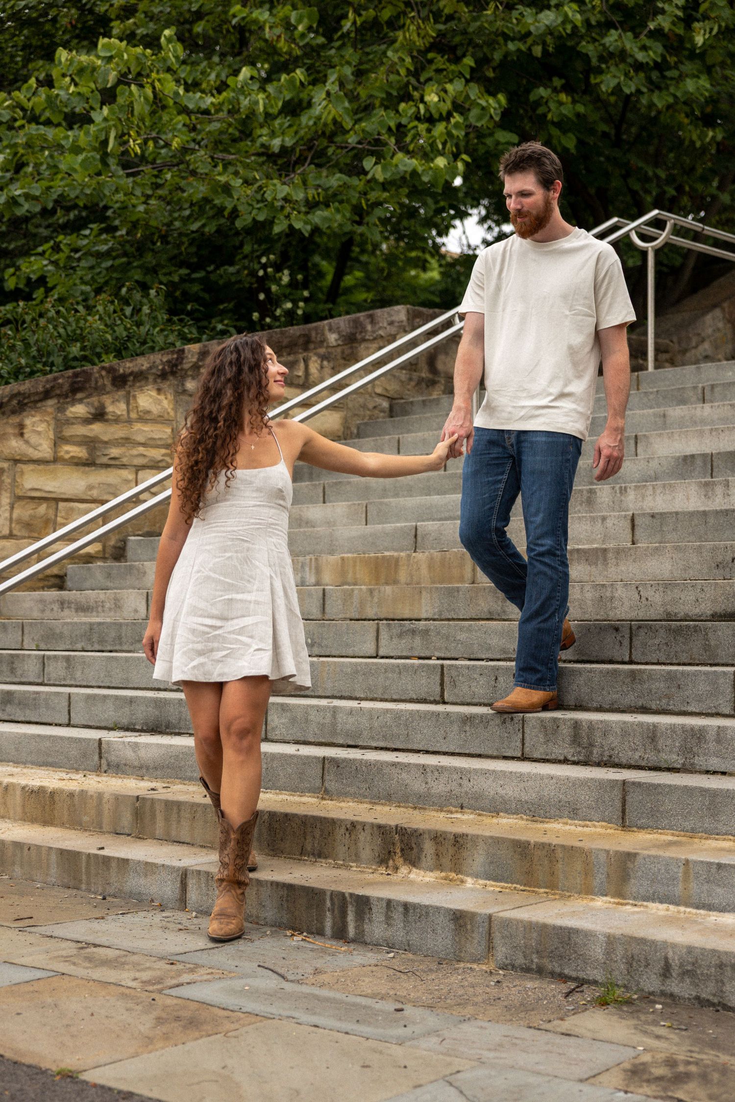 Couple holding hands on outdoor stone steps, woman in white dress and boots, man in jeans and white tee.