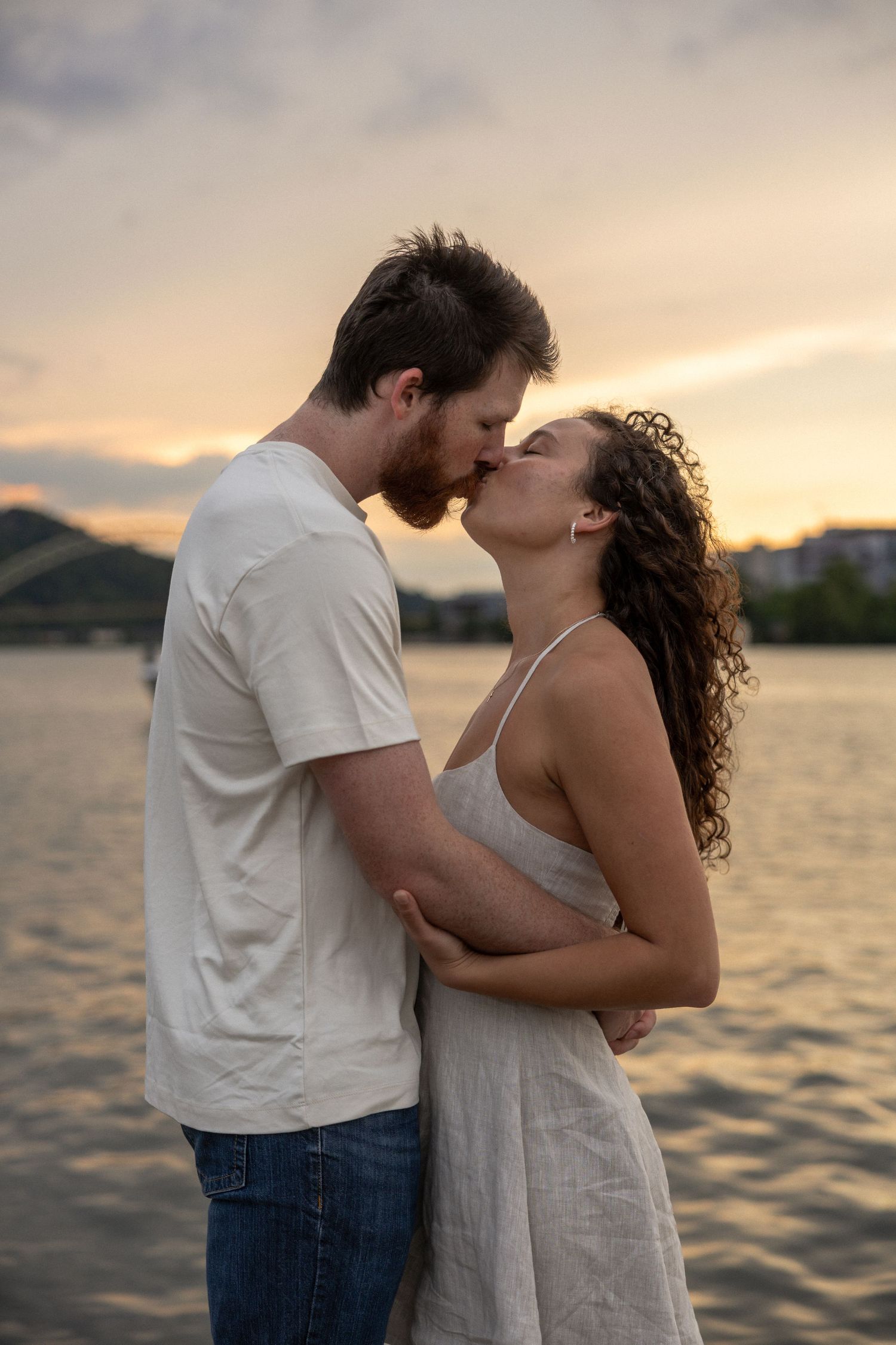A couple shares a romantic kiss by the water at sunset, with mountains and a glowing sky in the background.