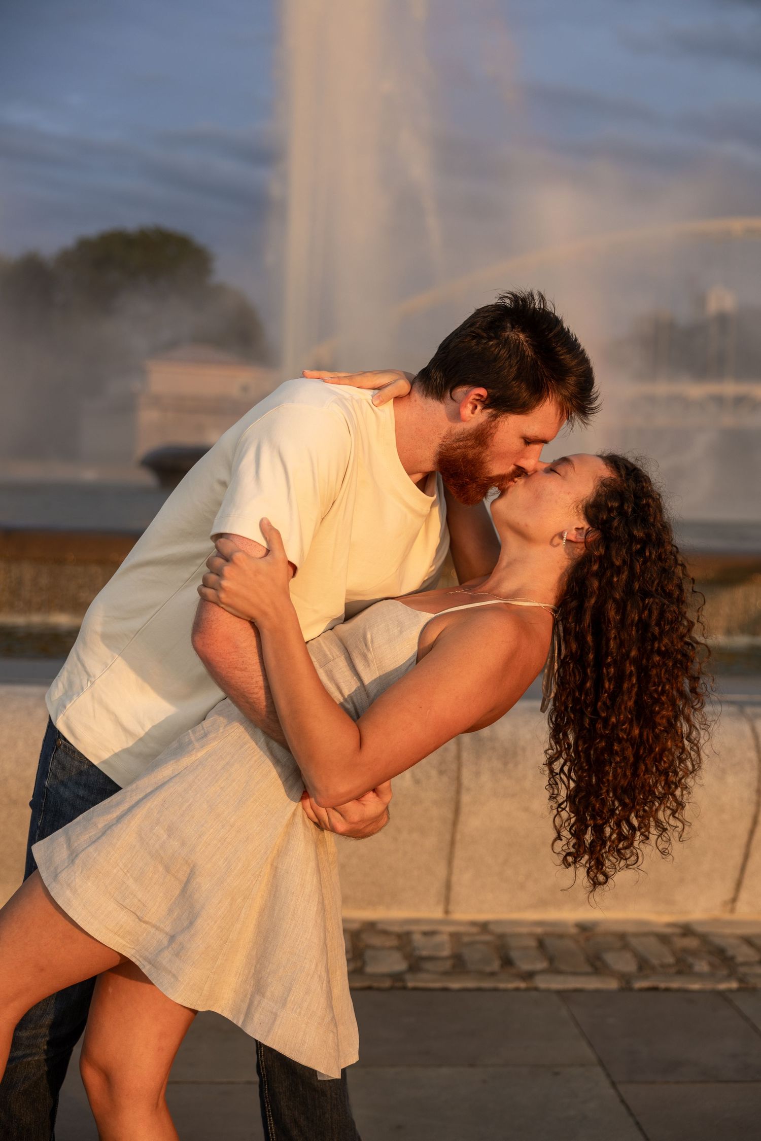 Couple sharing a romantic kiss near a fountain at golden hour, woman in white dress being dipped by man in white shirt.