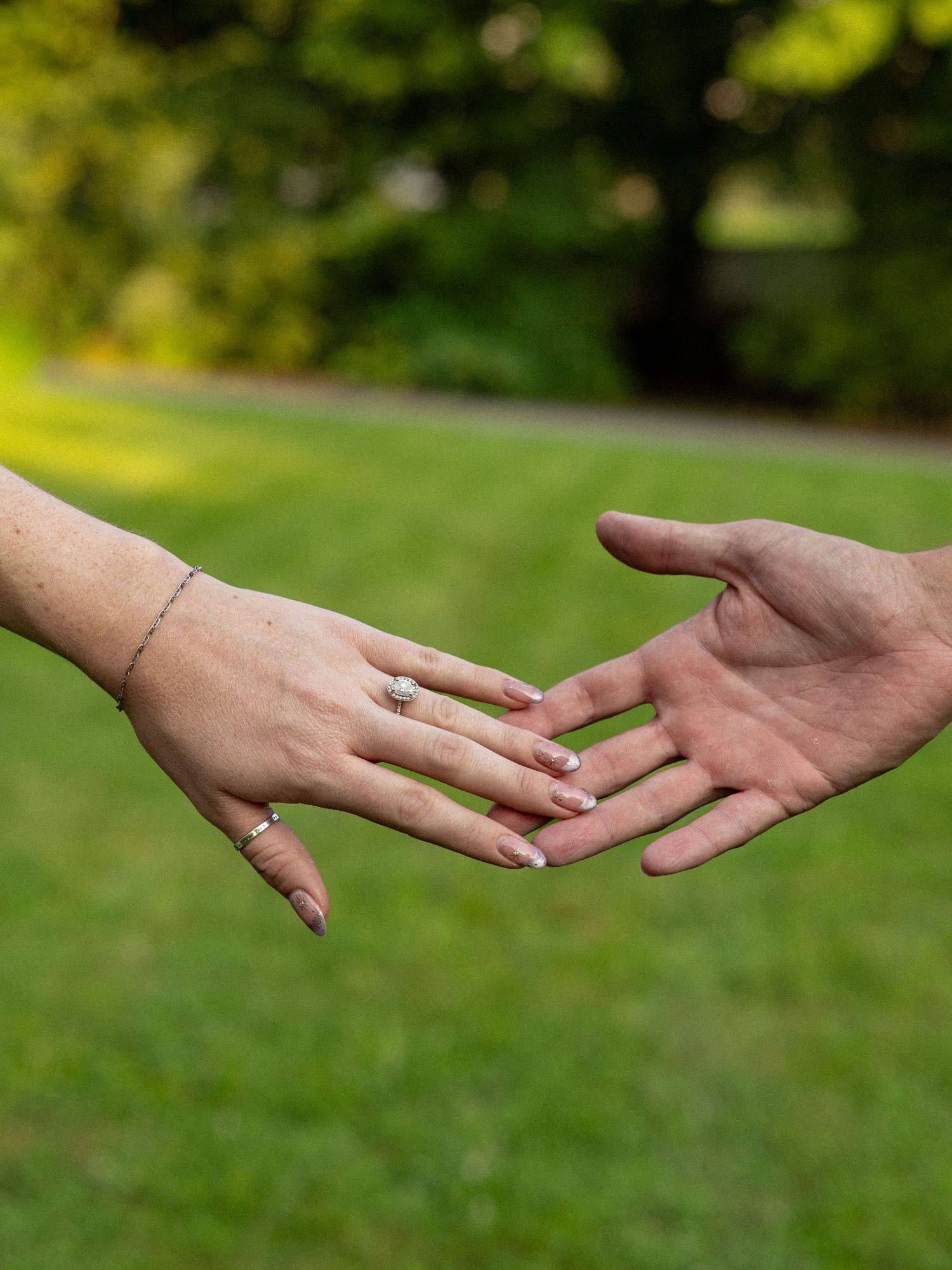 Two hands reaching toward each other outdoors, one wearing an engagement ring and bracelet, set against a green lawn.