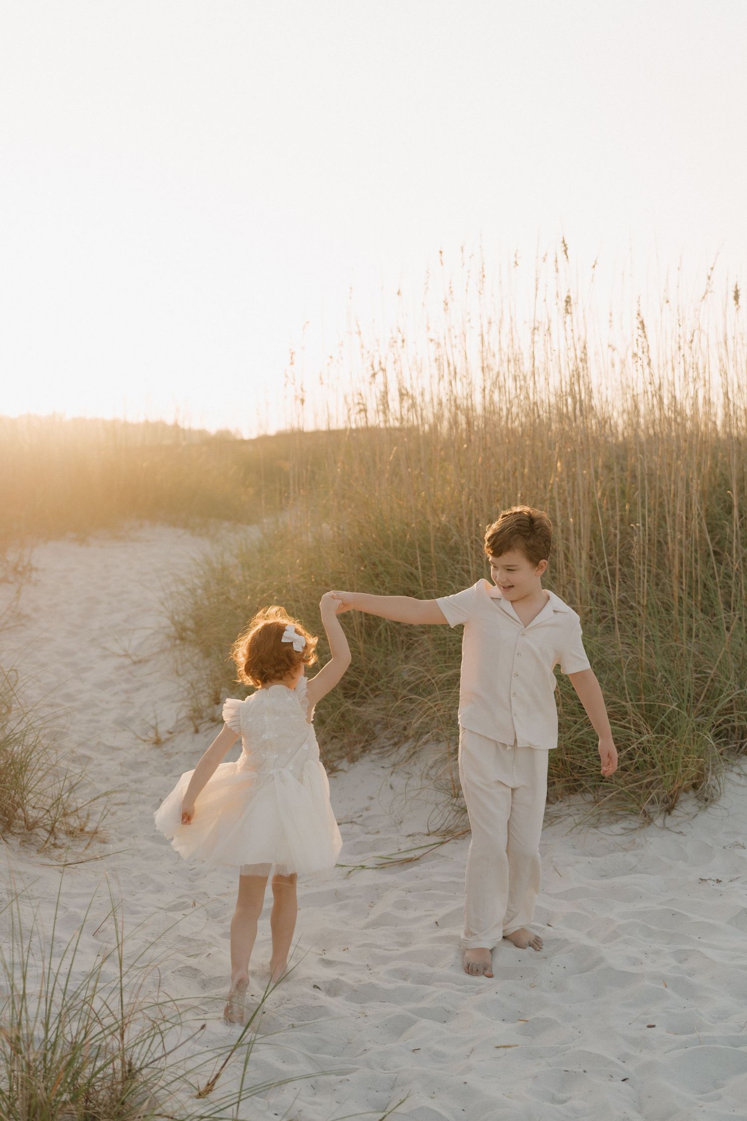 Children play and dance together on sandy beach dunes at sunset with tall grass in the background.
