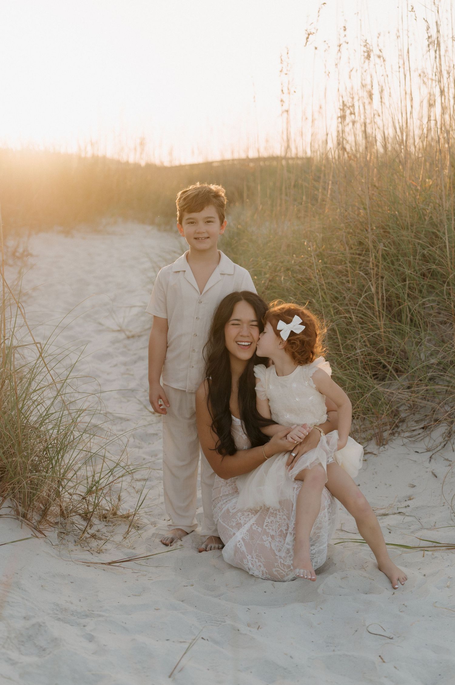 A couple sits together on a sandy beach at sunset with tall grass in the background.