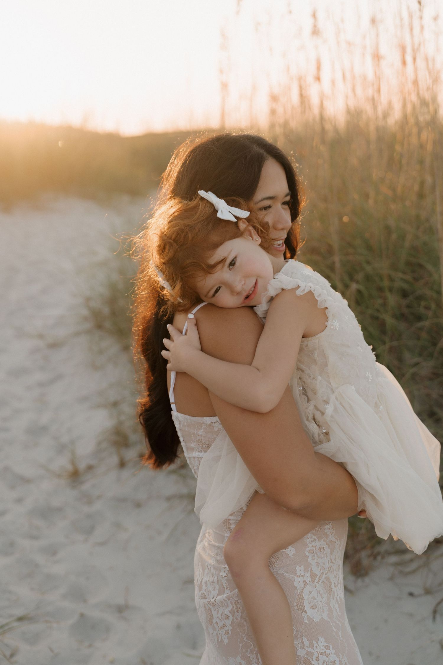 A tender embrace between two figures in white lace dresses on a sandy beach at sunset.