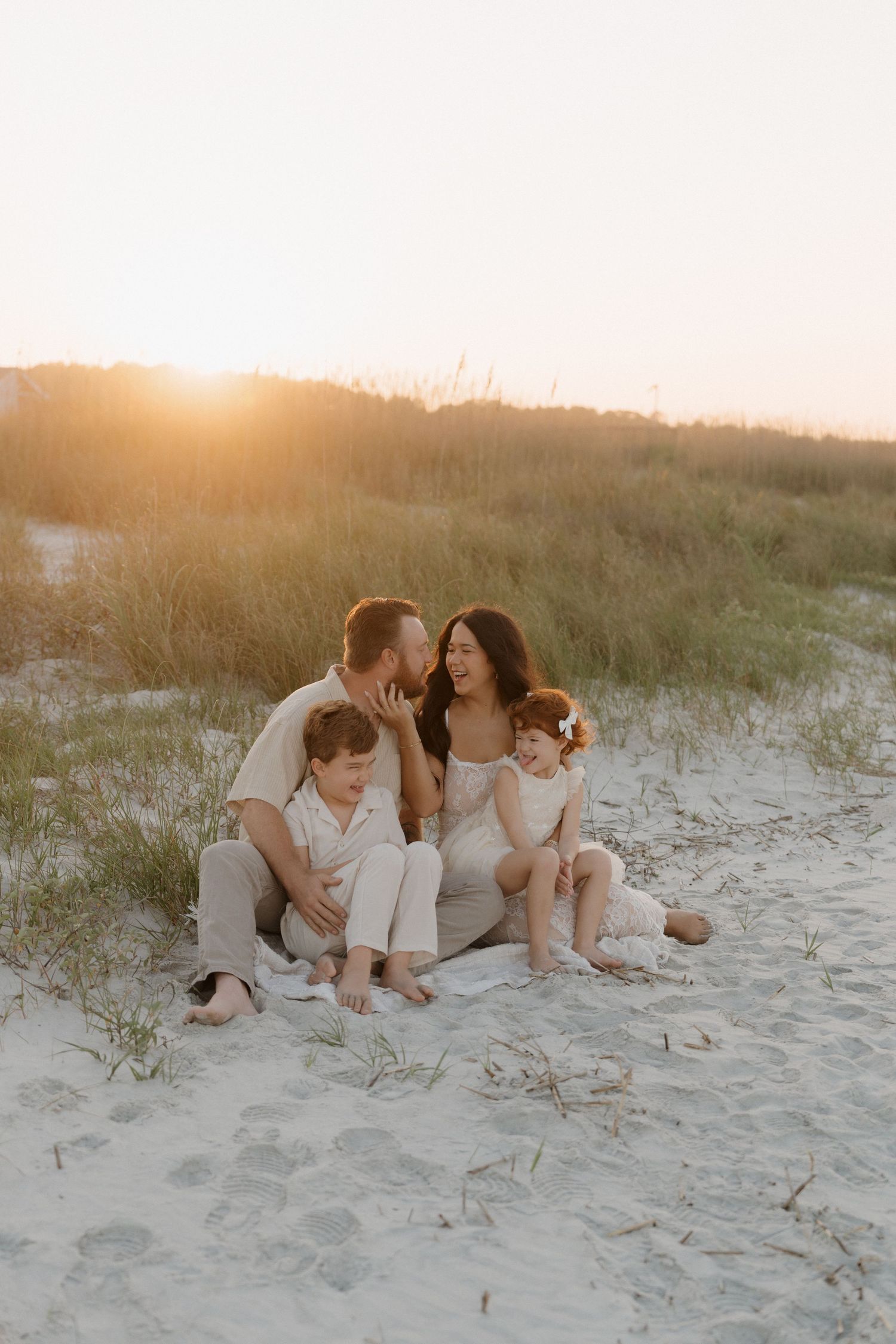 A family dressed in white sits together on a sandy beach dune during golden hour sunset.