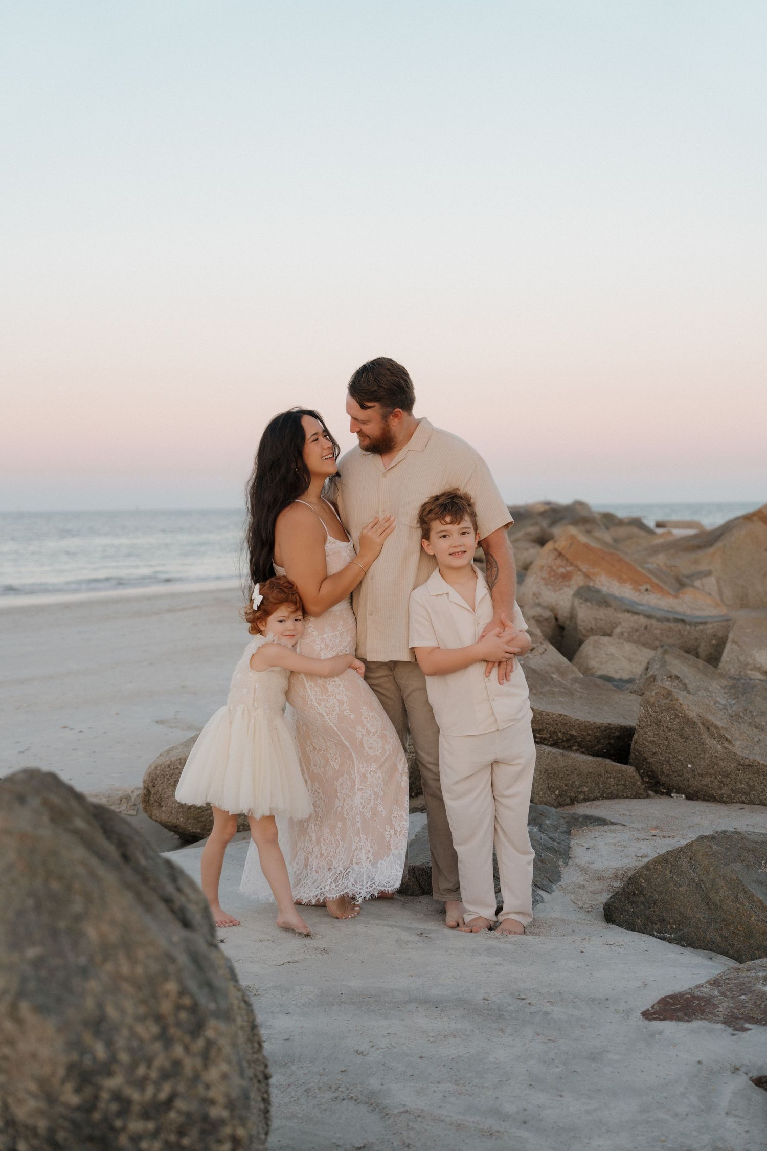A family embraces together on a rocky beach at sunset, wearing coordinated neutral-toned outfits.