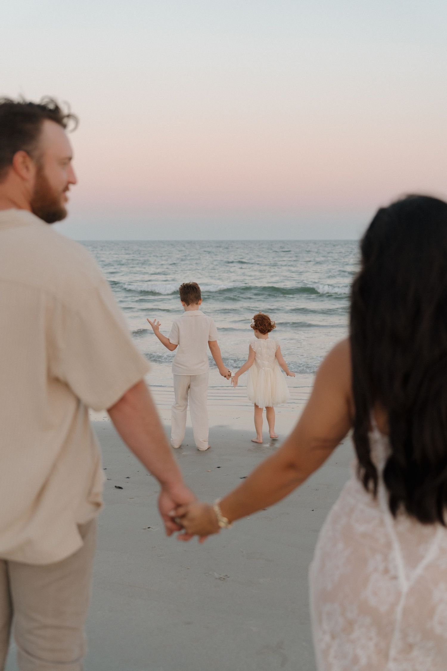 A couple holding hands walks along a peaceful beach at sunset, wearing light colored clothing.