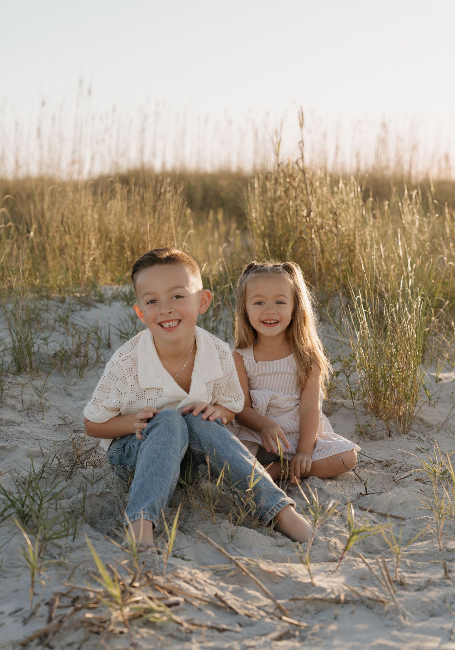 Two children sitting together on sandy beach dunes with tall grass during golden hour.