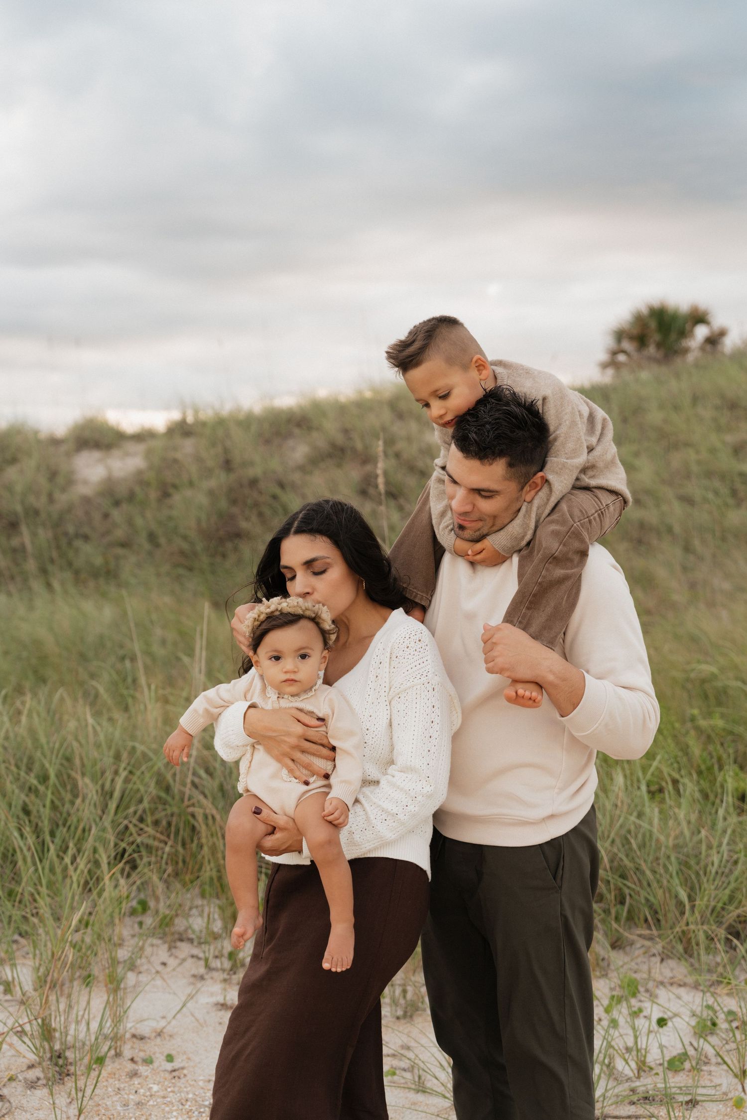 A loving family shares tender moments together on grassy sand dunes at sunset wearing coordinated neutral outfits.