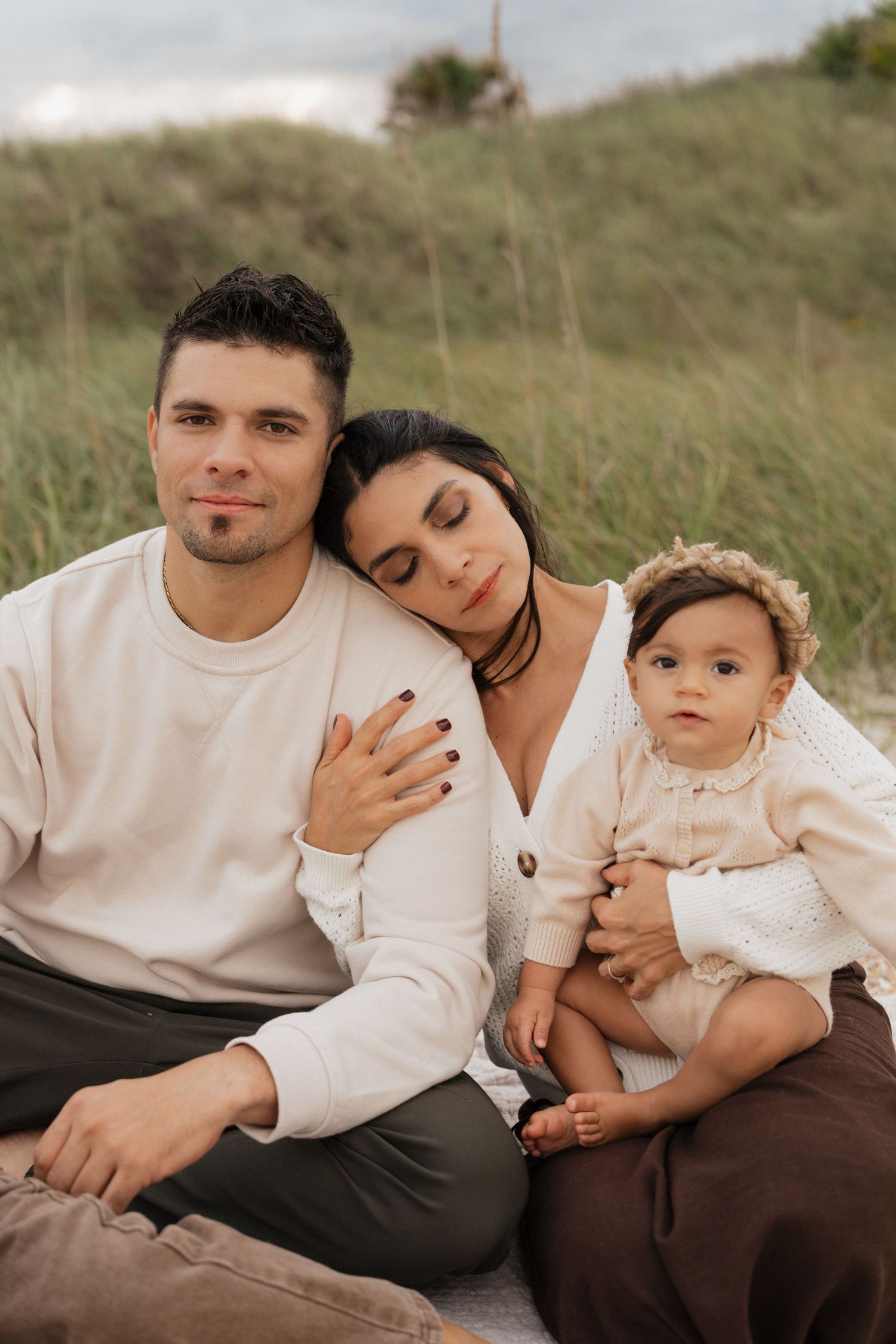 A family in white and brown outfits shares tender moments while sitting together in a grassy field.