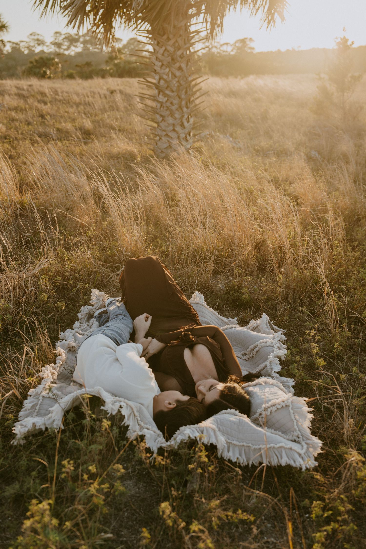A couple lies in tall golden grass during sunset with flowing white fabric creating a dreamy atmosphere.