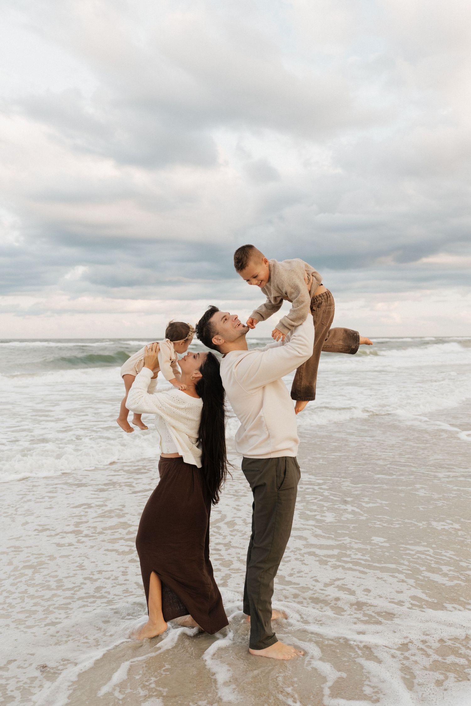 A family plays together on a cloudy beach, dipping and dancing in the ocean waves.